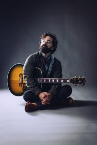 Andrew Duhon sits crosslegged with an acoustic guitar