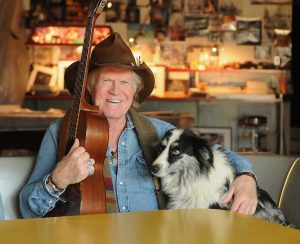 Billy Joe Shaver seated with his acoustic guitar and a black and white dog