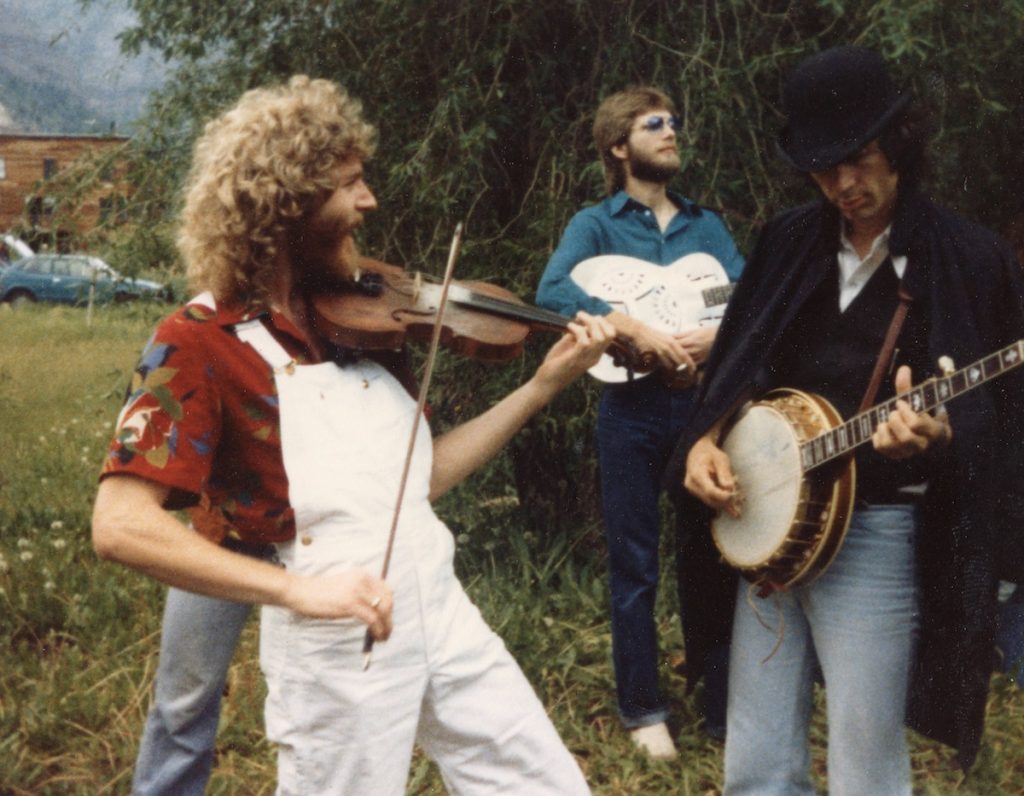 Sam Bush plays fiddle and John Hartford plays banjo in a grassy area