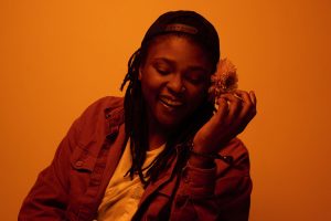 Joy Oladokun with a backward ballcap and a flower behind her ear, washed in orange light