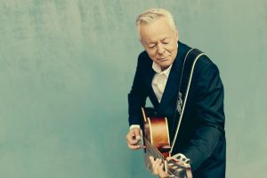 Tommy Emmanuel in blue suit with acoustic guitar against a sea green background
