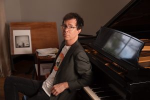 Ben Folds sits on a piano bench with his back to the keys