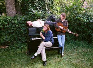 The three members of Bonny Doon posed around a piano on a green lawn