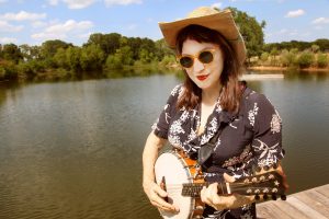 Ann Savoy by a lake, holding a banjo and wearing sunglasses